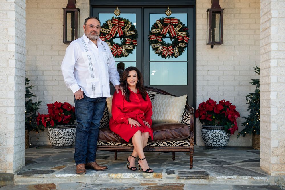 Couple posing on a home's front porch decorated for Christmas with red bow wreaths on the door and poinsettias in planters.