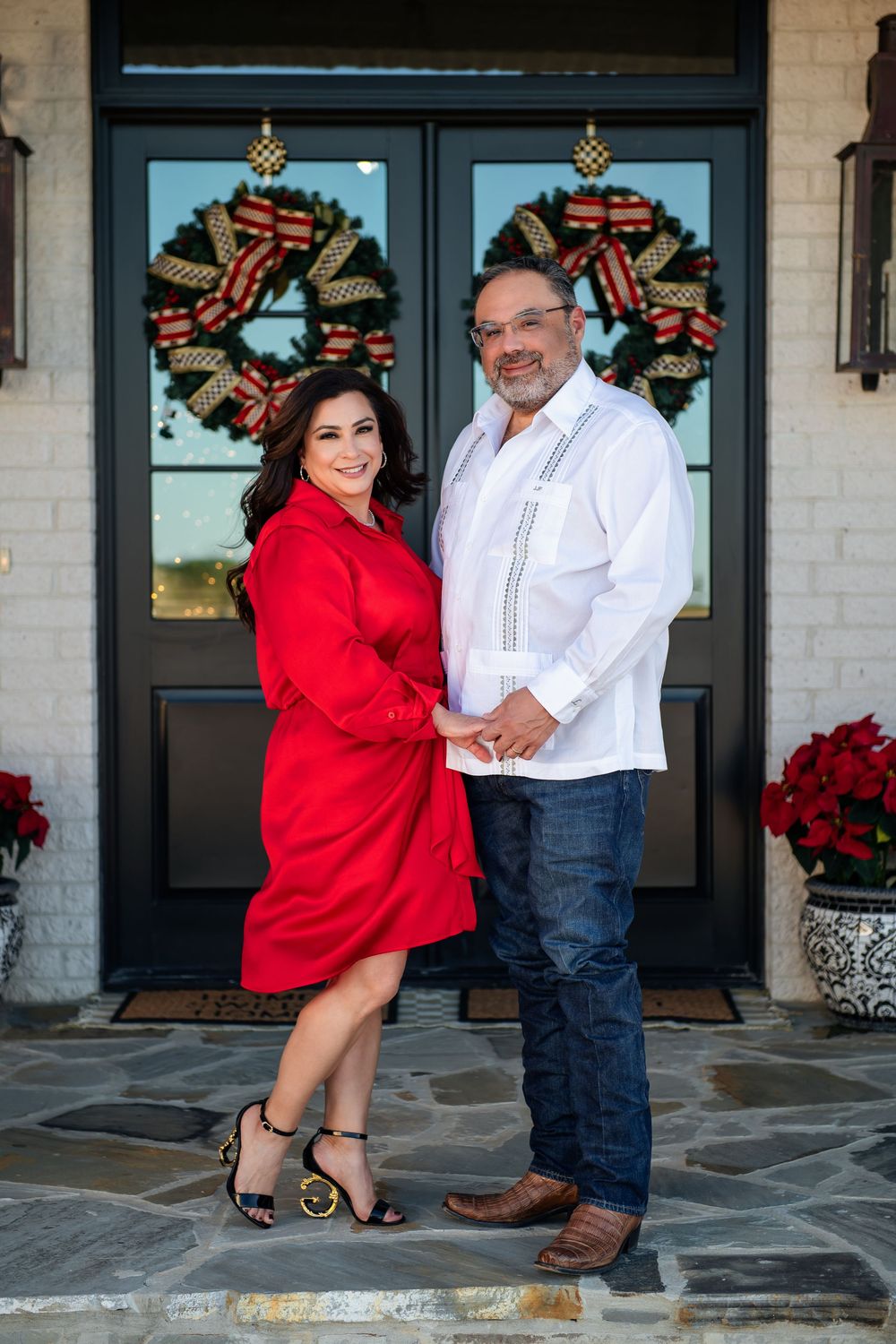 Smiling couple in matching holiday outfits hold hands on a stone porch in front of a door with red bow wreaths.