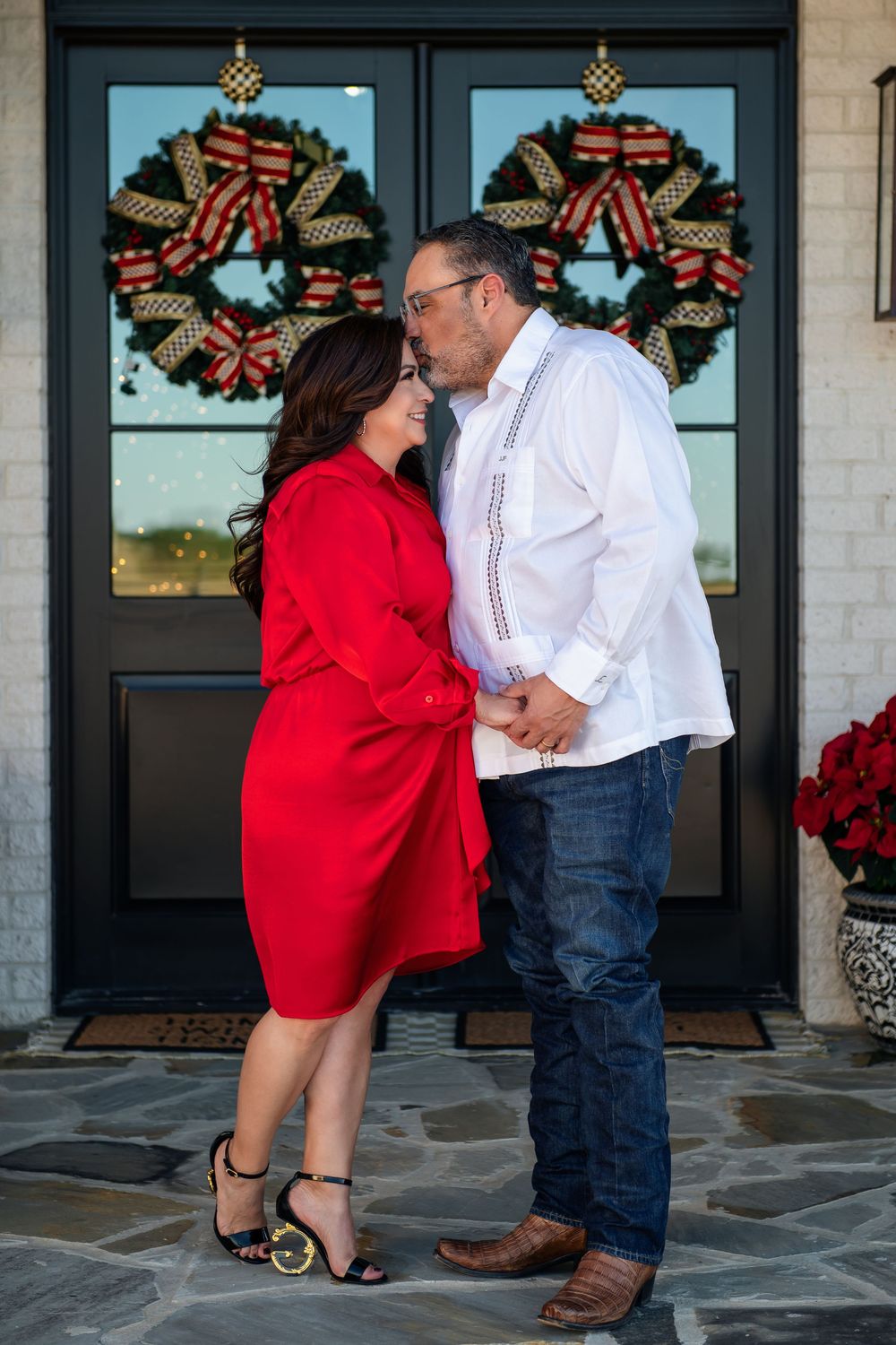 Couple in red dress and white shirt share a kiss on a stone porch beneath festive Christmas wreaths.