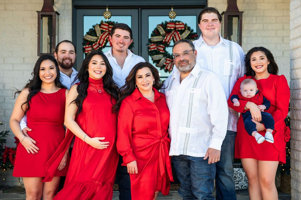 Multigenerational family dressed in red and white gathers close for a Christmas portrait outside a brick home.