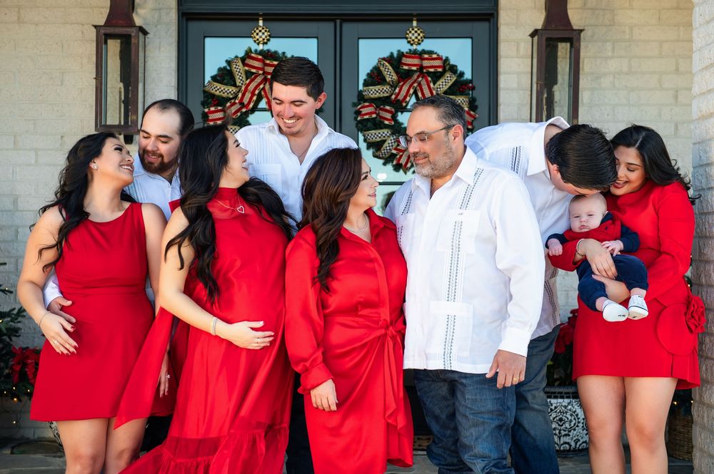 Family in red and white outfits gathers on a porch with holiday wreaths, with one man holding a baby.