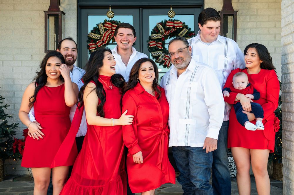 A large family in red and white outfits, including a baby, gathers on a stone porch decorated with Christmas wreaths.
