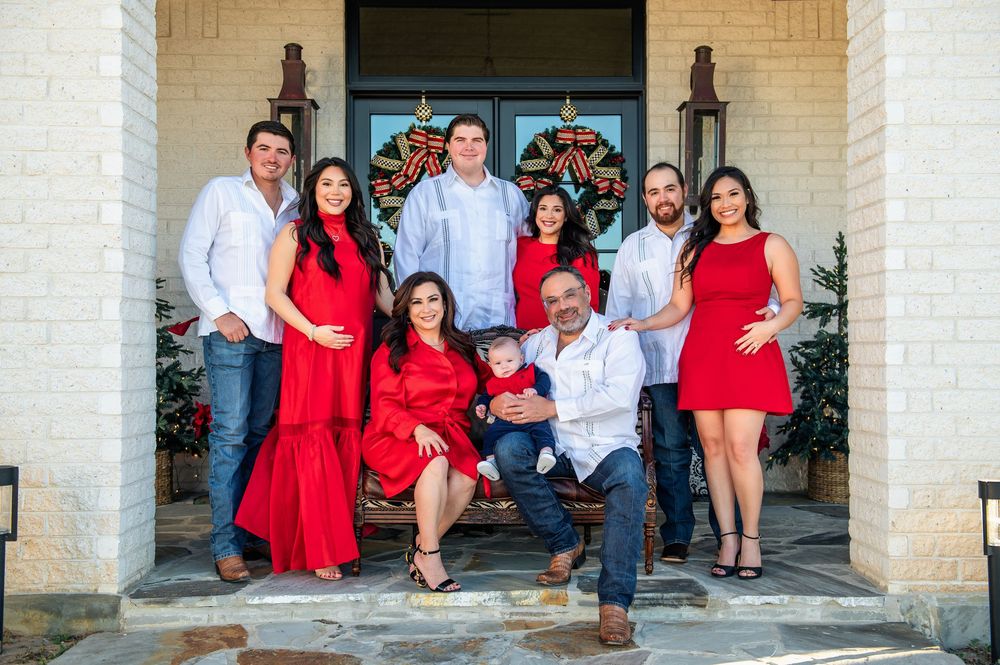 Family of nine in red and white outfits poses on the front steps of a brick home decorated with Christmas wreaths.