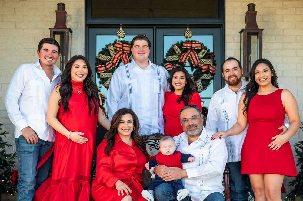 A multigenerational family in red and white poses together on a front porch with holiday wreaths while grandparents hold a baby.