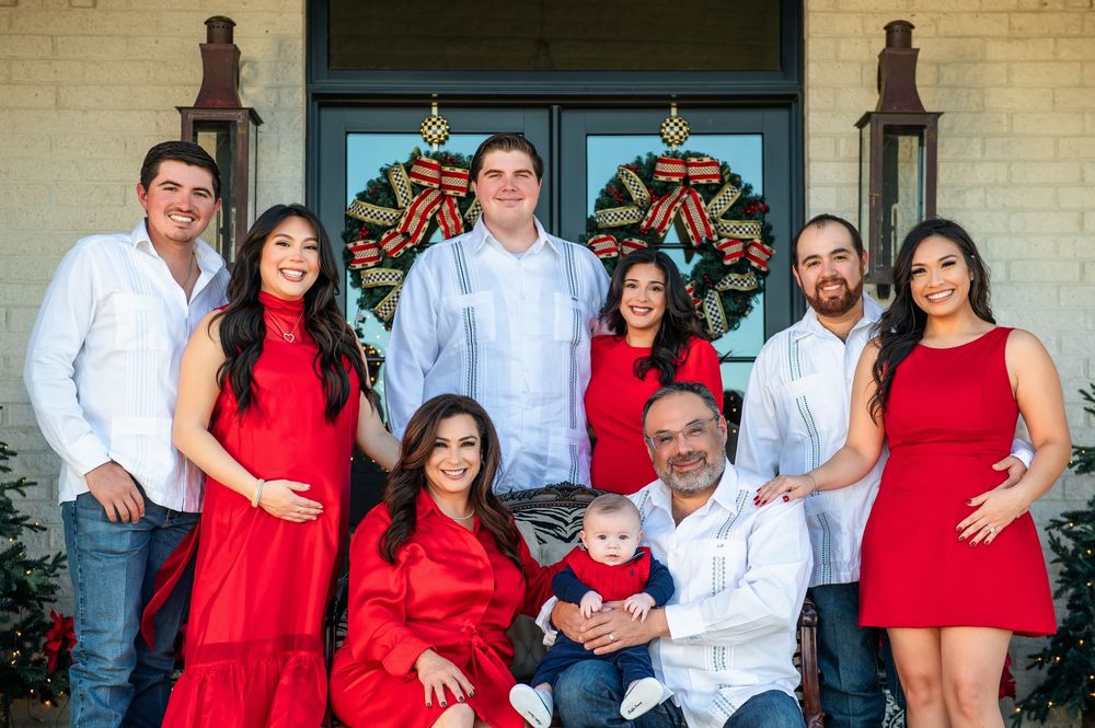 A joyful extended family dressed in red and white smiles on a brick home's porch while grandparents hold a baby in front of wreaths.