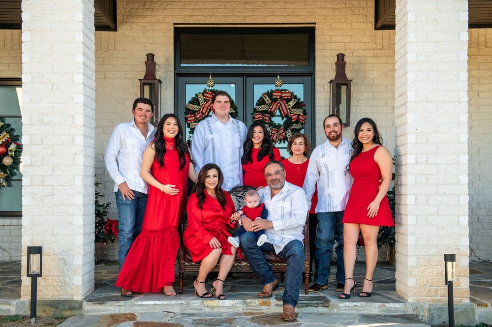 Large family in red and white outfits gathers on a brick home porch decorated with Christmas wreaths.
