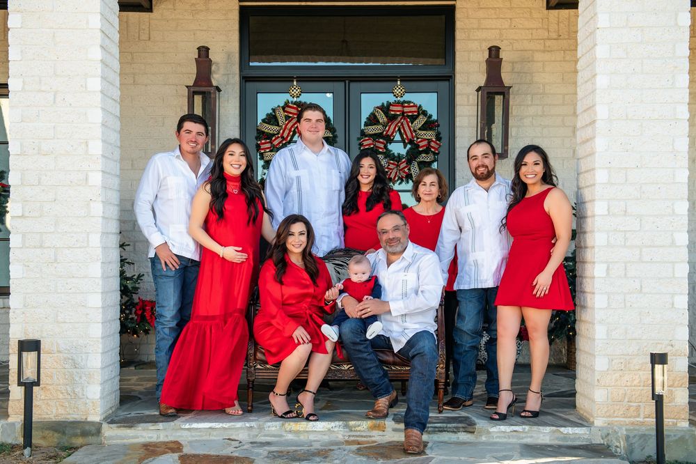 A full family portrait in red and white attire on the steps of a brick home with Christmas wreaths on the front doors.