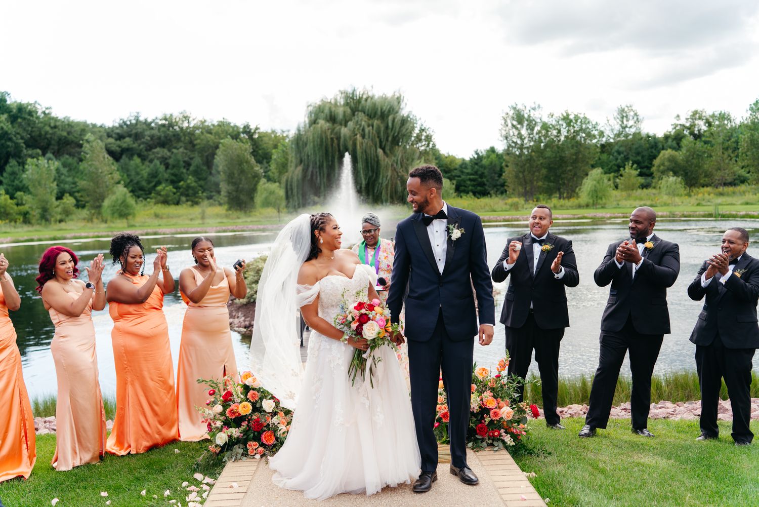 Bride and groom walk down an aisle past cheering bridesmaids in coral dresses and groomsmen in dark suits at a lakeside outdoor wedding.