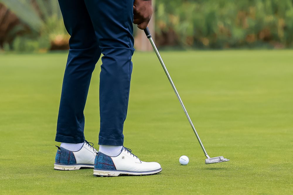 Golfer in navy pants and blue and white golf shoes lines up a putt with a putter beside a golf ball on a green.
