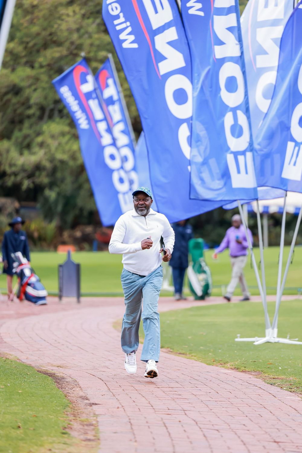 Man in a white jacket, jeans and cap jogs along a brick path at a golf event lined with blue sponsor flags and spectators.