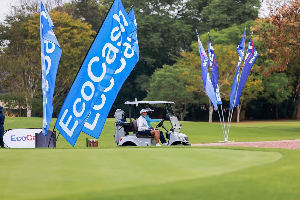 Golfer seated in a white golf cart on a green fairway beside blue EcoCash sponsor flags at a tournament surrounded by trees.