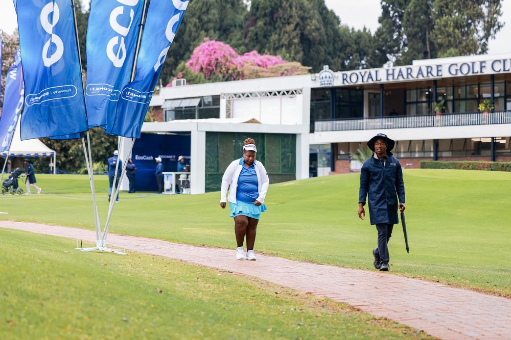 Two golfers walk along a paved path past blue flags toward the Royal Harare Golf Club clubhouse on a green course.