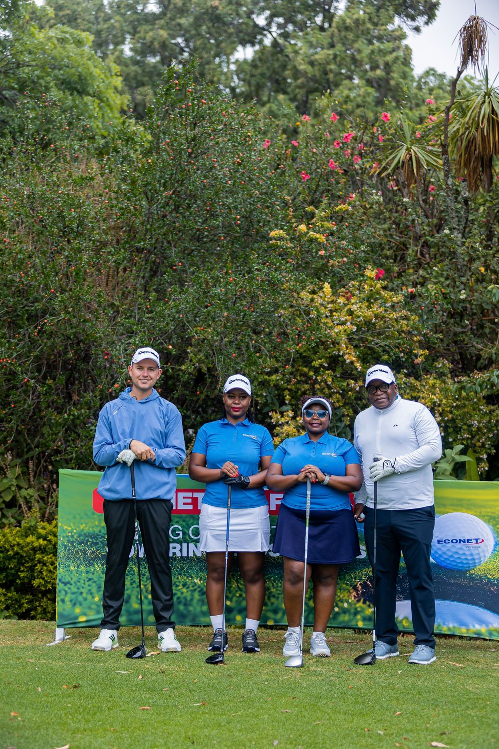 Four golfers in matching caps and polos pose with clubs on a lush green course in front of a golf tournament sponsor banner.