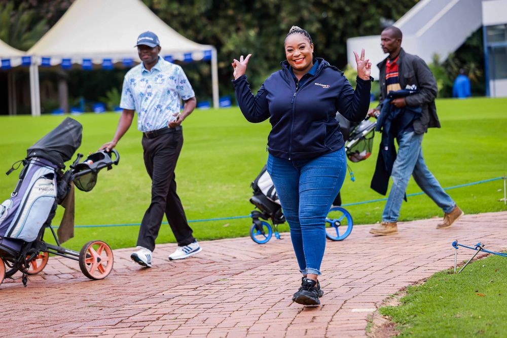 Smiling woman in a navy jacket and jeans waves cheerfully on a brick path at a golf course while two men walk behind with golf trolleys.