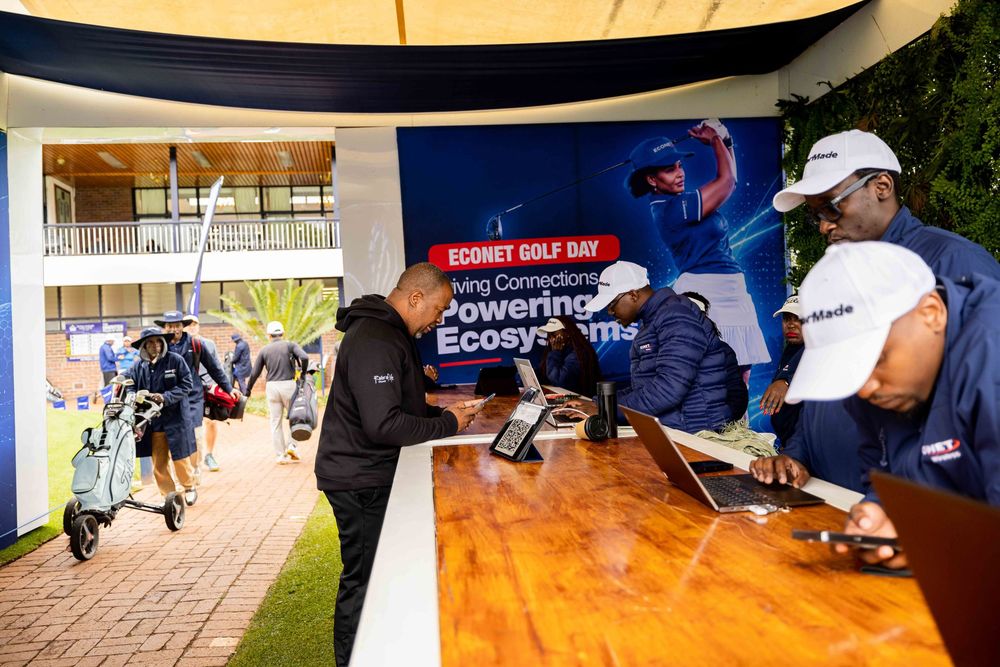 Staff and guests in caps work at laptops on a wooden counter at an Econet Golf Day registration desk beside a golf cart and course path.