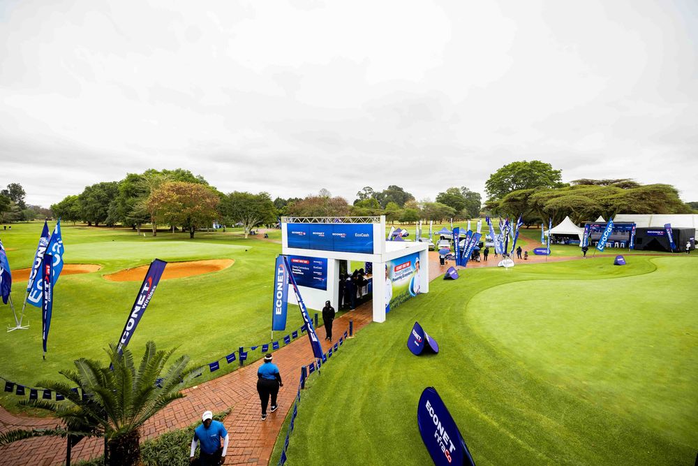 Blue tournament banners and a hospitality pavilion line a brick pathway beside a lush golf course green as spectators walk toward the event.