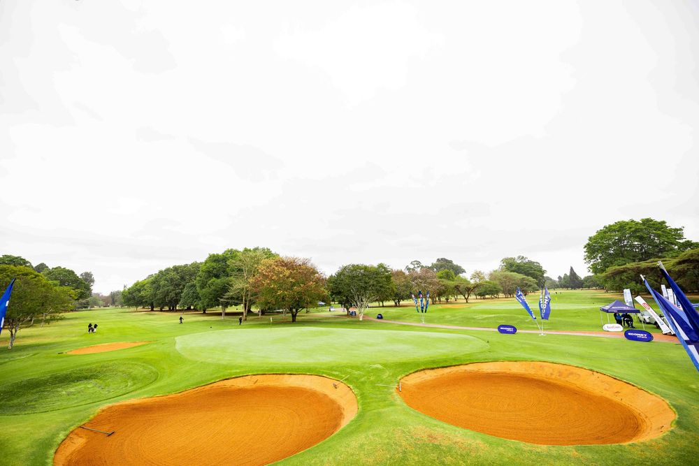 Golf course green with two orange sand bunkers in the foreground, tournament banners, and mature trees under an overcast sky.