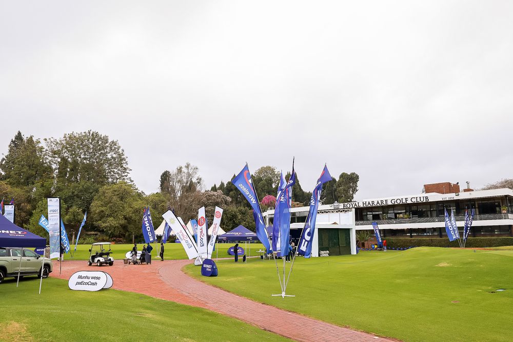 Blue promotional flags and tents line a red brick path beside a golf club clubhouse on a green course under an overcast sky.