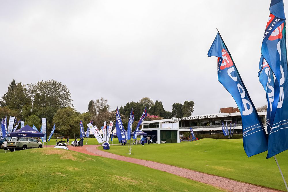 Blue promotional flags and event tents line a golf course fairway beside a clubhouse with trees and an overcast sky in the background.