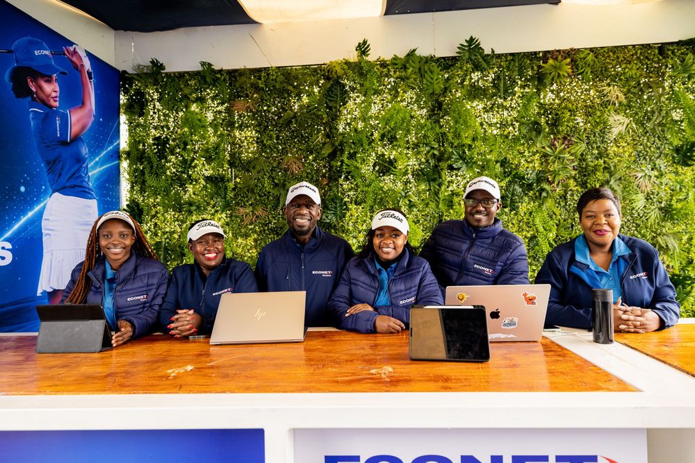 Six smiling staff members in navy jackets and white caps sit behind a wooden counter with laptops in front of a green plant wall.