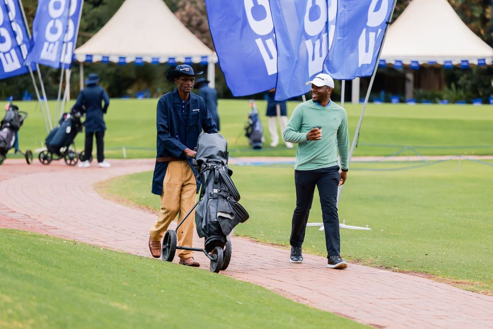 Two golfers walk along a brick cart path pulling push carts past blue banners and white tents on a green golf course.