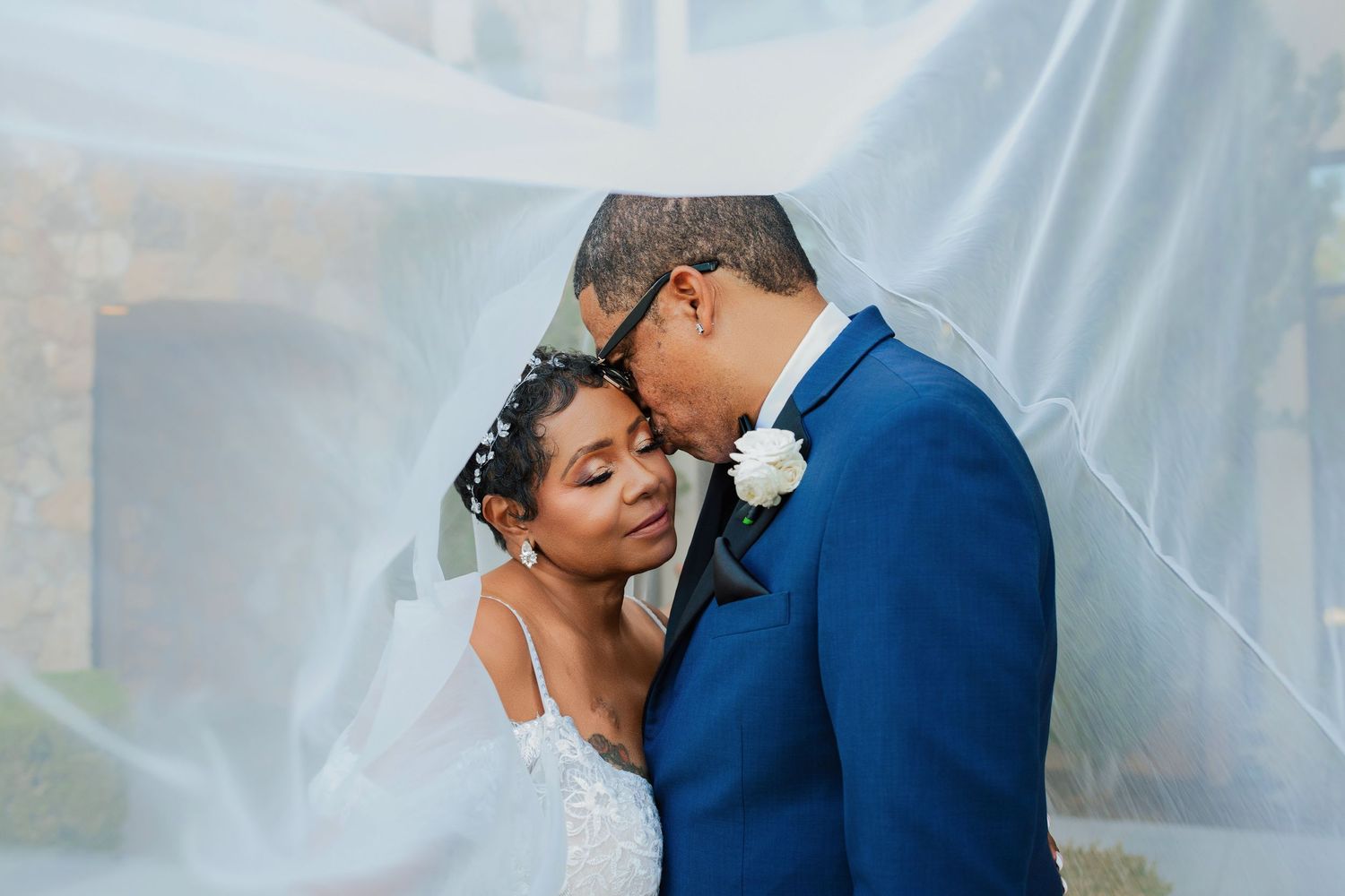 Bride in a white lace gown and groom in a blue suit with a white boutonniere embrace tenderly beneath a sheer veil.