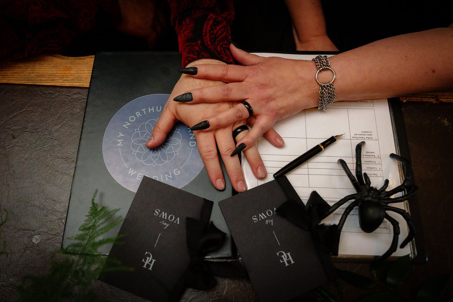 Couple's hands with black nails and silver rings signing the wedding register beside a black spider decoration