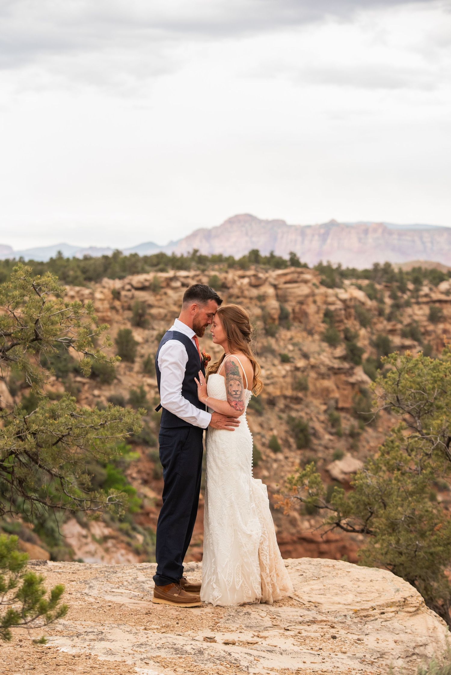Bride and groom embrace on a sandstone cliff edge during an elopement with desert canyon mesas and juniper trees in the background.