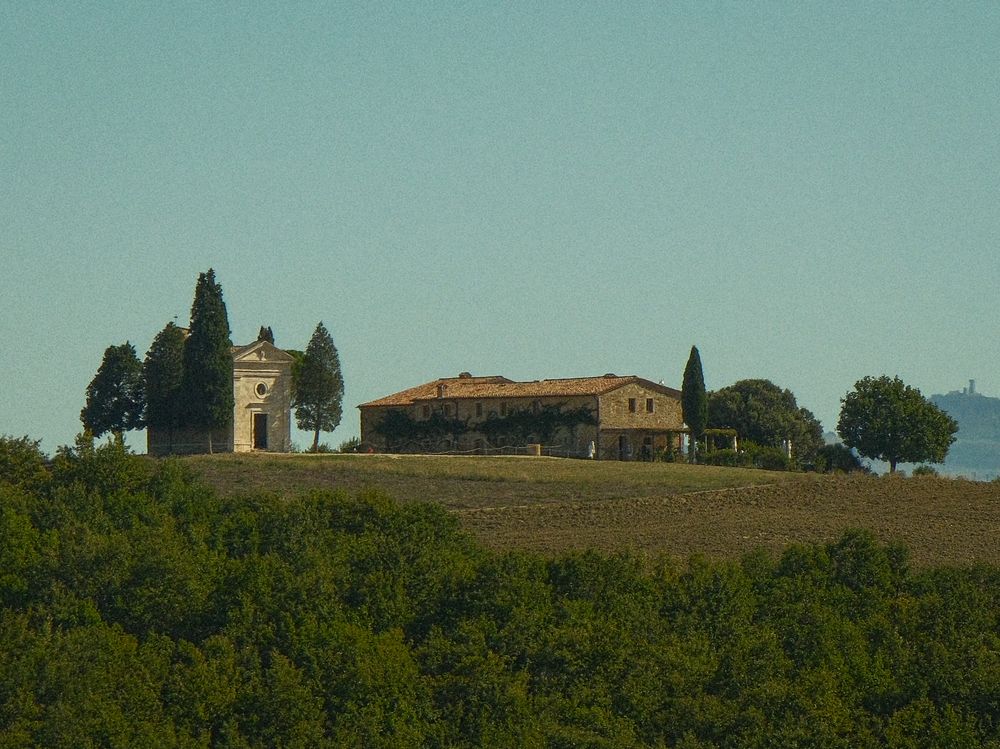 Rustic Tuscan stone farmhouse and small chapel framed by tall cypress trees on a grassy hilltop above green vineyards under a clear sky.