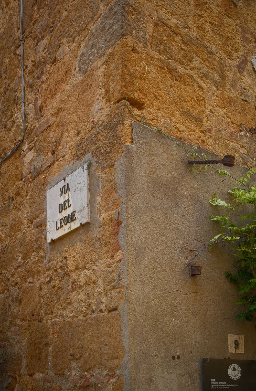 Weathered white street sign reading Via del Leone on a rustic golden stone building corner in a historic Italian village.