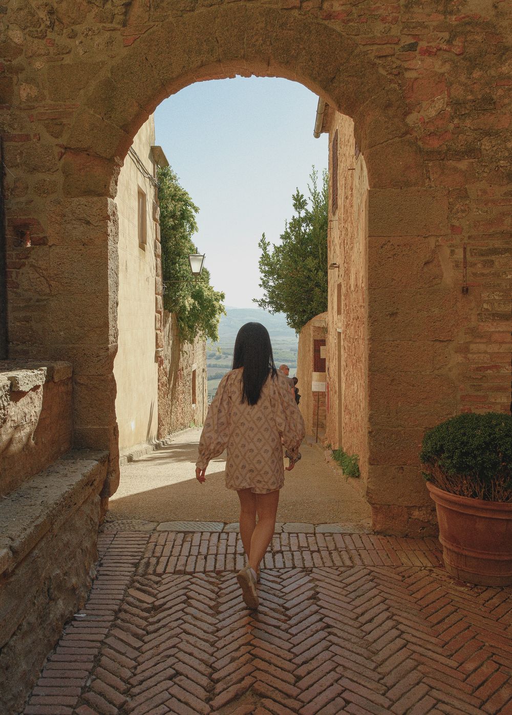 Woman in a floral dress walks through a sunlit stone archway on a herringbone brick path toward a scenic Tuscan hillside view.