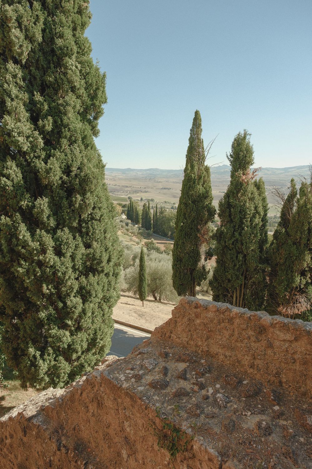 Tall cypress trees beside a weathered stone wall overlook a sunlit Tuscan valley with rolling hills under a clear blue sky.