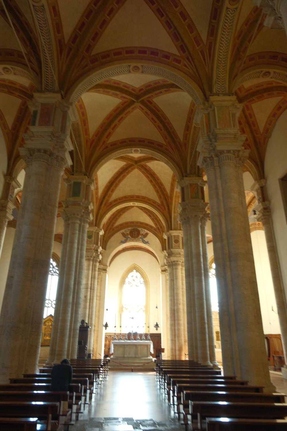 Gothic church interior with tall stone columns, ribbed vaulted ceilings with painted details, wooden pews and a lit stained glass window.