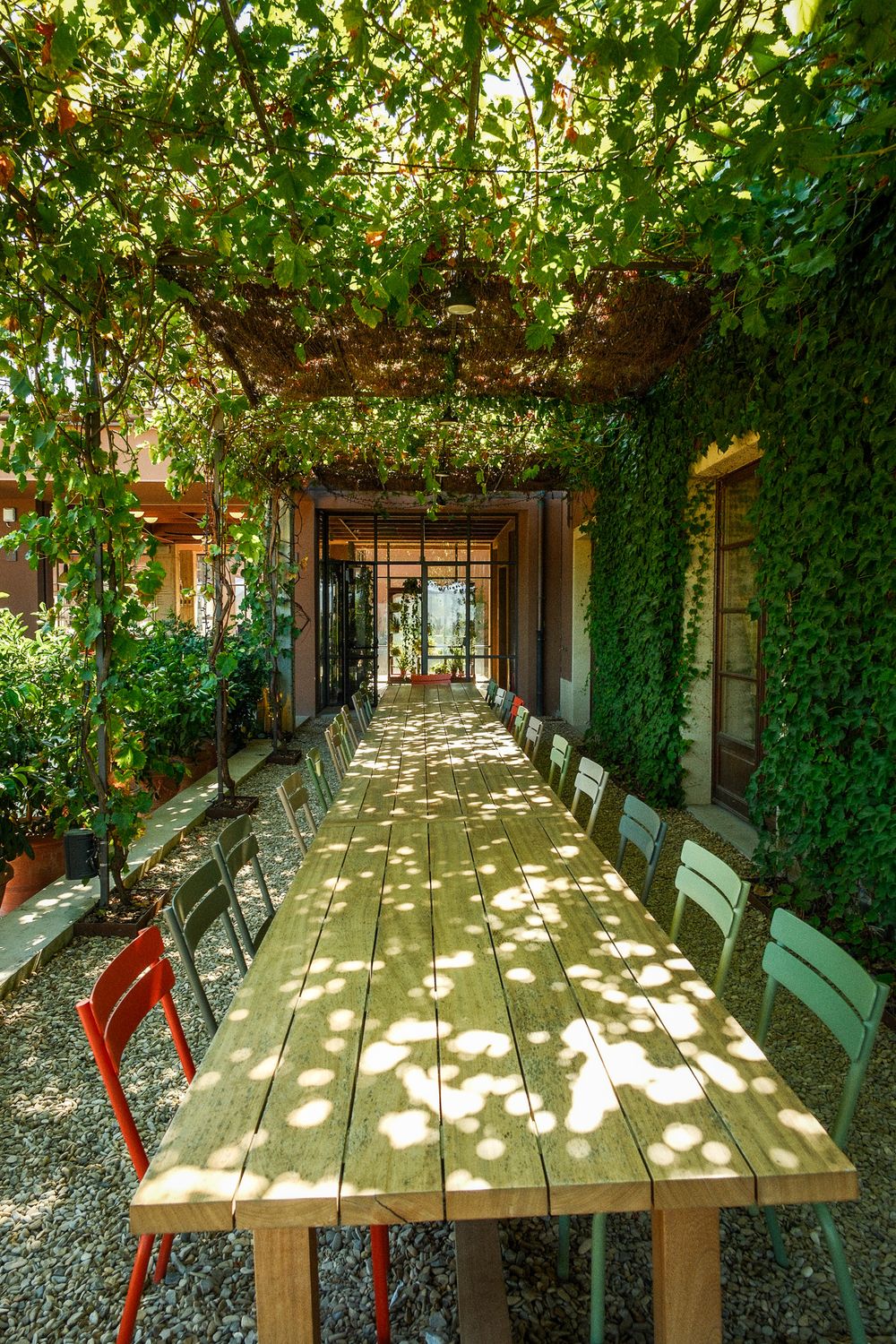 Long wooden outdoor dining table with colorful chairs under a vine-covered pergola, dappled sunlight and a gravel patio.