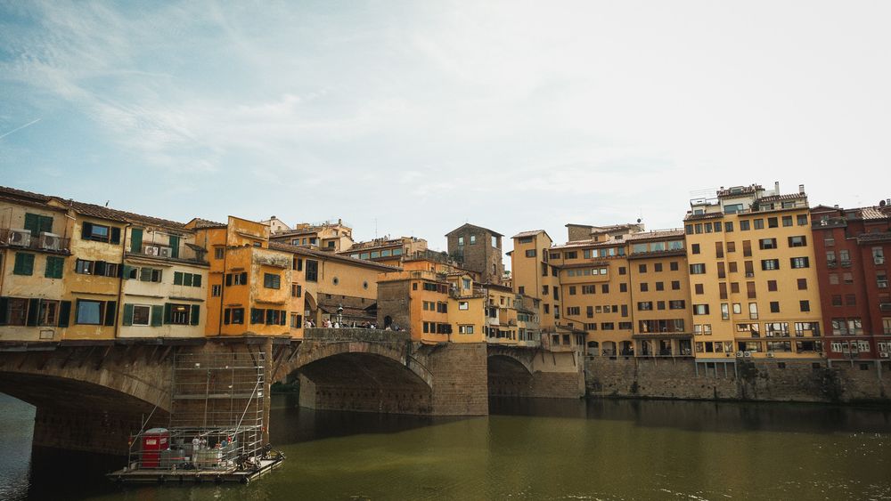 Ponte Vecchio bridge in Florence, Italy, with colorful medieval buildings reflected in the calm Arno River under a soft blue sky.