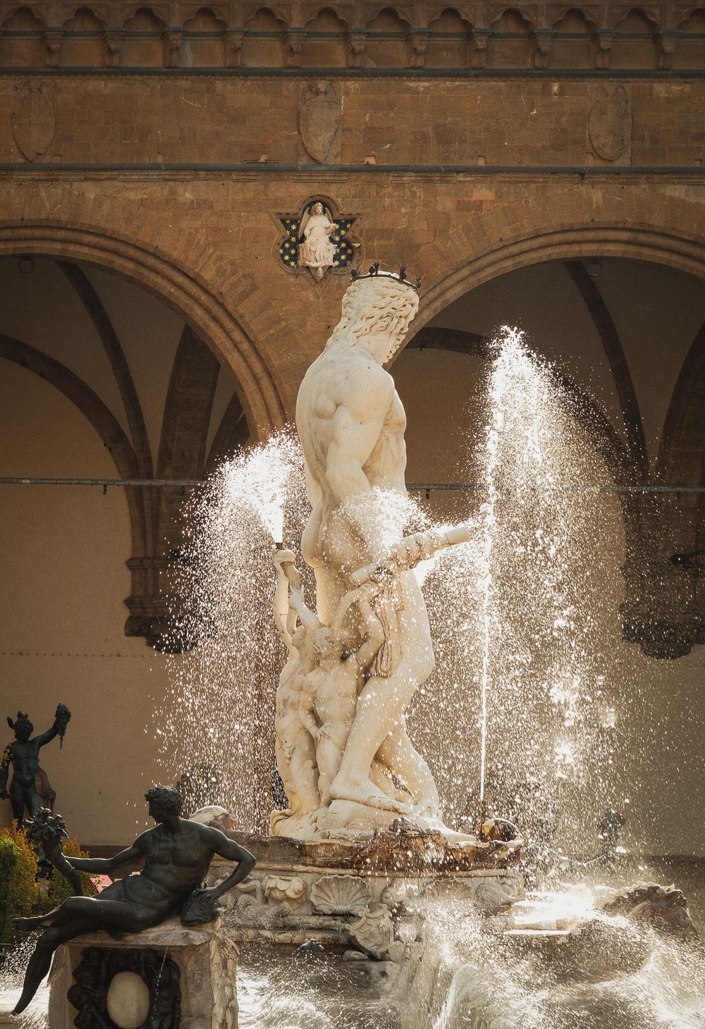 Marble statue of Neptune rising above splashing water in the Fountain of Neptune in Florence, with a bronze satyr figure in the foreground.