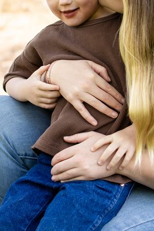 A mother with blonde hair gently holds her young child in a brown ribbed top on her lap outdoors.
