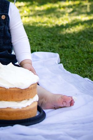 Baby sitting on white blanket outdoors beside a layered cake with white frosting on a black stand.