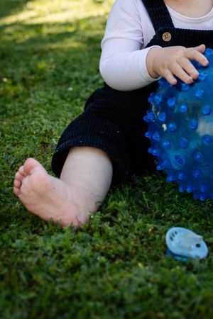 Baby sitting on green grass outdoors, holding a blue spiky sensory ball, wearing dark overalls and white shirt.
