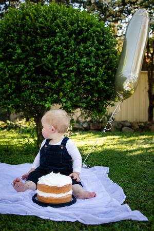 Baby in overalls sits on white blanket outdoors with smash cake and silver balloon for first birthday celebration.
