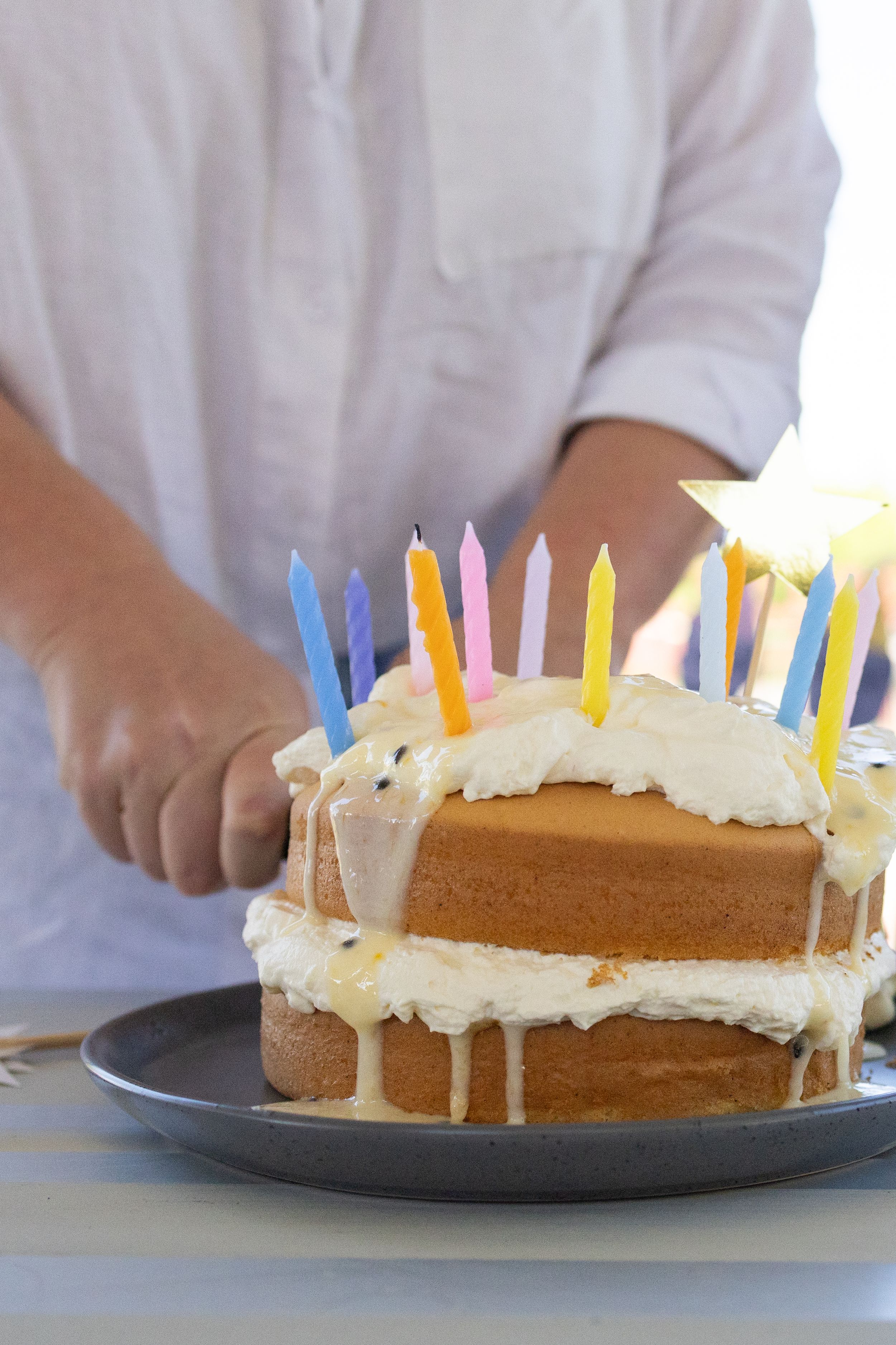 Person placing colorful birthday candles on a two-layer white frosted cake on a gray plate.
