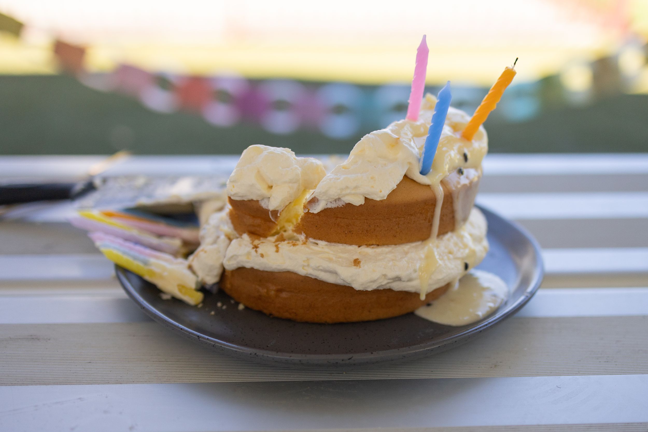 A birthday cake with white frosting and colorful candles on a gray plate outdoors on a white wooden table.