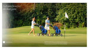 Two golfers walking on a lush green golf course pulling blue golf bags on carts on a sunny day.