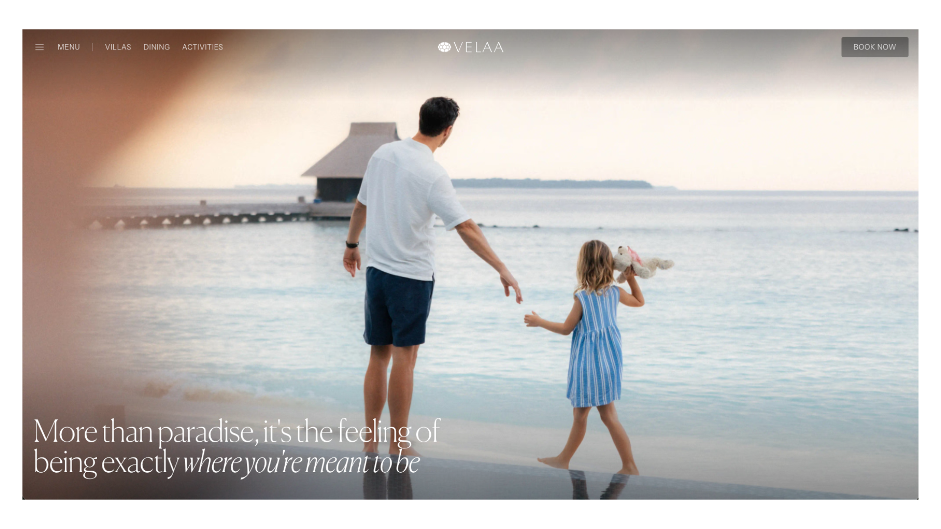 A father and young daughter hold hands on a tropical beach at sunset, with an overwater bungalow in the distance.