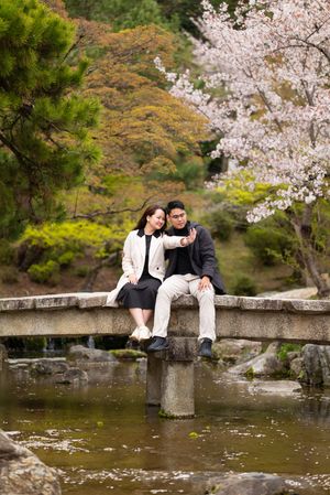 Couple taking a selfie on a stone bridge in a Japanese garden with cherry blossoms in bloom.