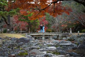 Couple standing on a stone bridge surrounded by vibrant autumn foliage in a Japanese garden.
