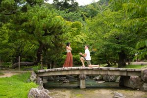 A couple shares a romantic proposal moment on a stone bridge surrounded by lush green trees in a Japanese garden.
