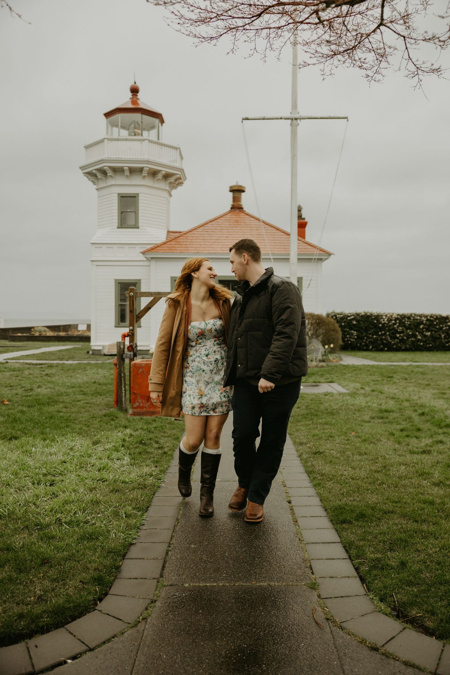 Couple walking hand in hand on a brick path in front of a white lighthouse on a cloudy day in Mukilteo