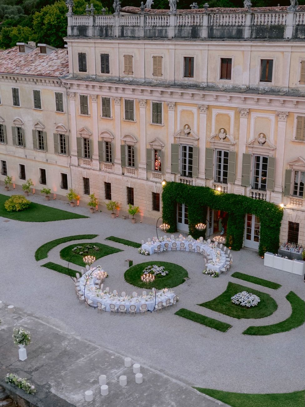 Wedding dinner setup with semi circular tables enlightened by glass chandeliers, in the garden of Villa Bettoni. Design Solidea Events