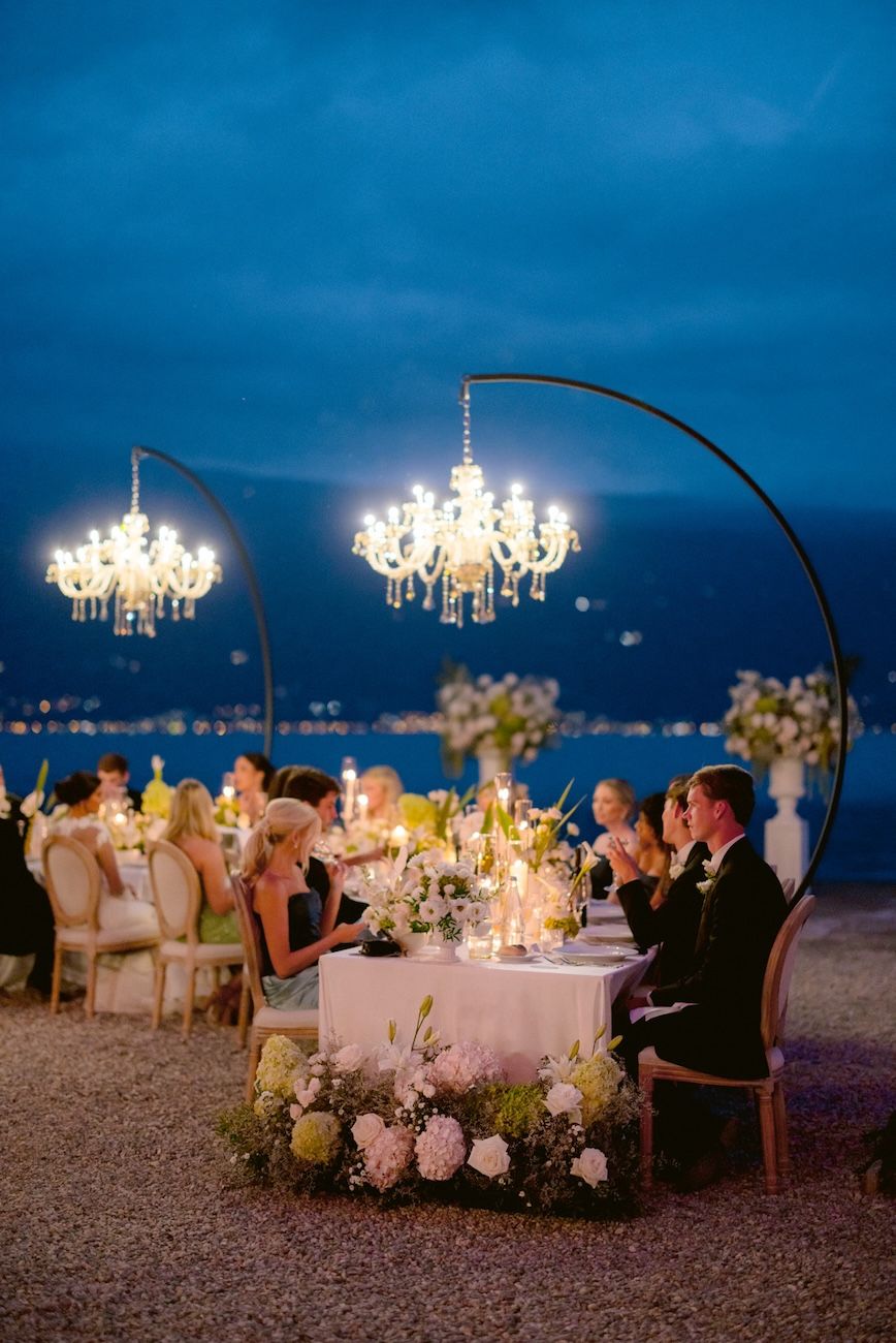 Wedding dinner setup with white, pale pink and green flowers on semi circular tables enlightened by glass chandeliers. Design Solidea Events